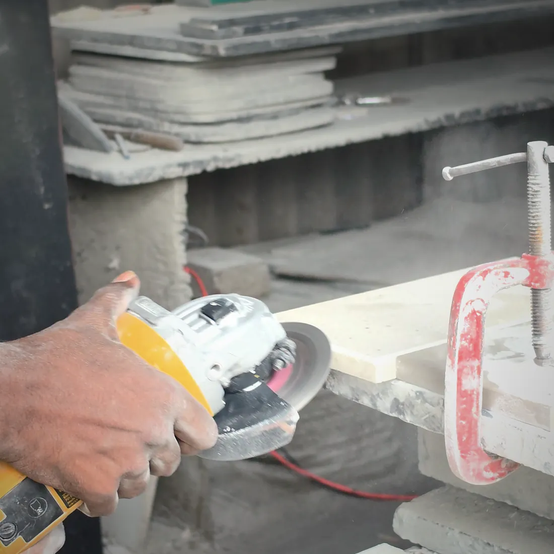 A hand guiding an angle grinder along the edge of a pale stone slab, dust rising from the cut.